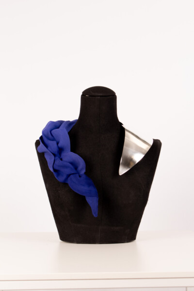 Mustalla mallinukella on oikealla puolella hopeinen metallinen olkaosa ja vasemmalla olkapäällä on punottu kuninkaansininen kangas, joka on raaputettu vasemmalle olkapäälle valkoista taustaa vasten.