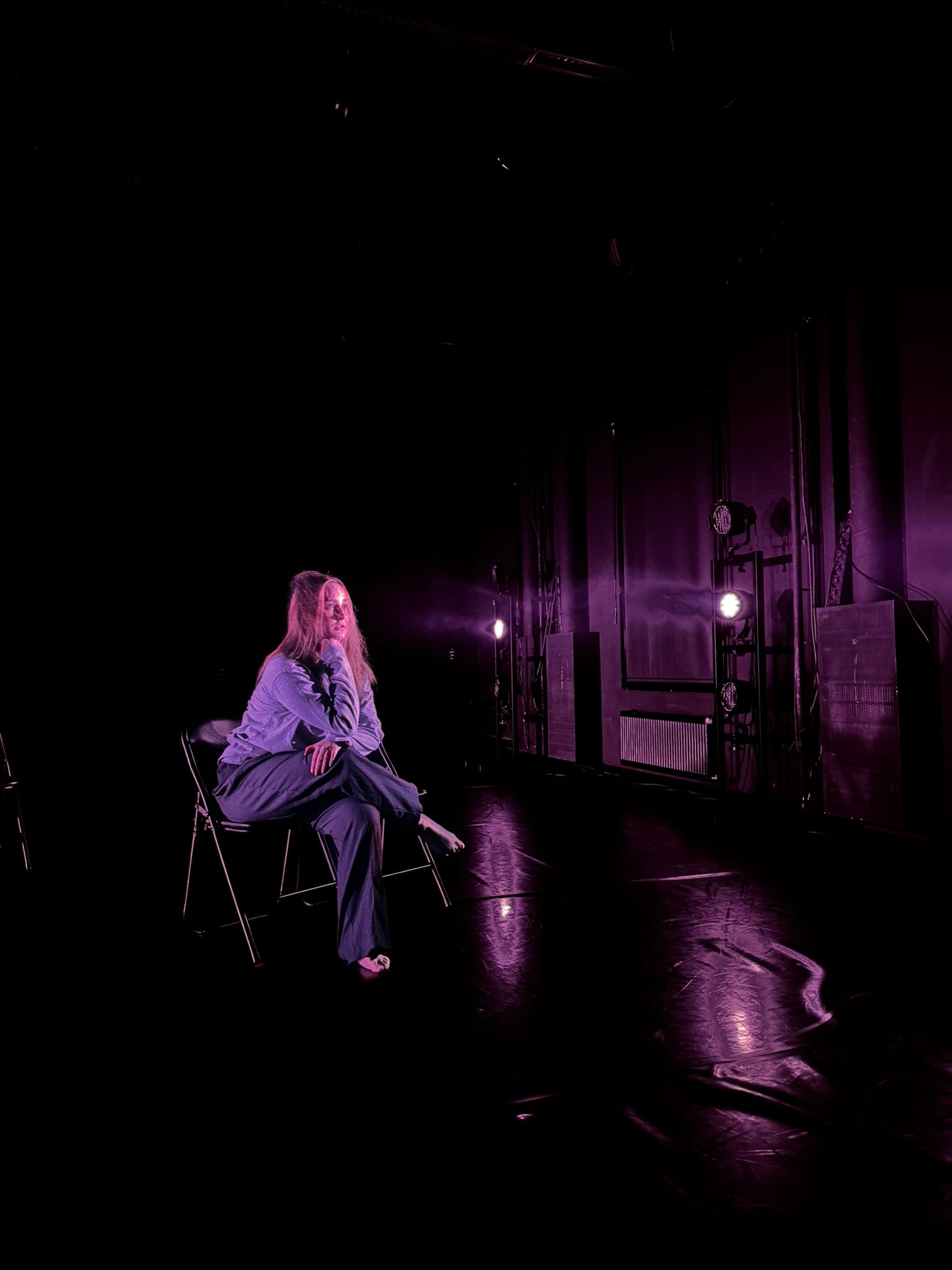 Pitkätukkainen nainen istuu yksin tuolilla pimeässä huoneessa, jota valaisevat violetit näyttämövalot. Tunnelma on tunnelmallinen ja dramaattinen, ja valo heijastuu kiiltävästä lattiasta.
