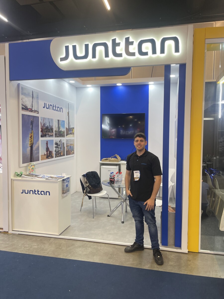 Mies seisoo Junttanin messuosaston edessä, jonka seinillä on valokuvia rakennuskoneista, televisioruutu ja tuoleja, ja osastolla on pöytä, jossa on vesipulloja.