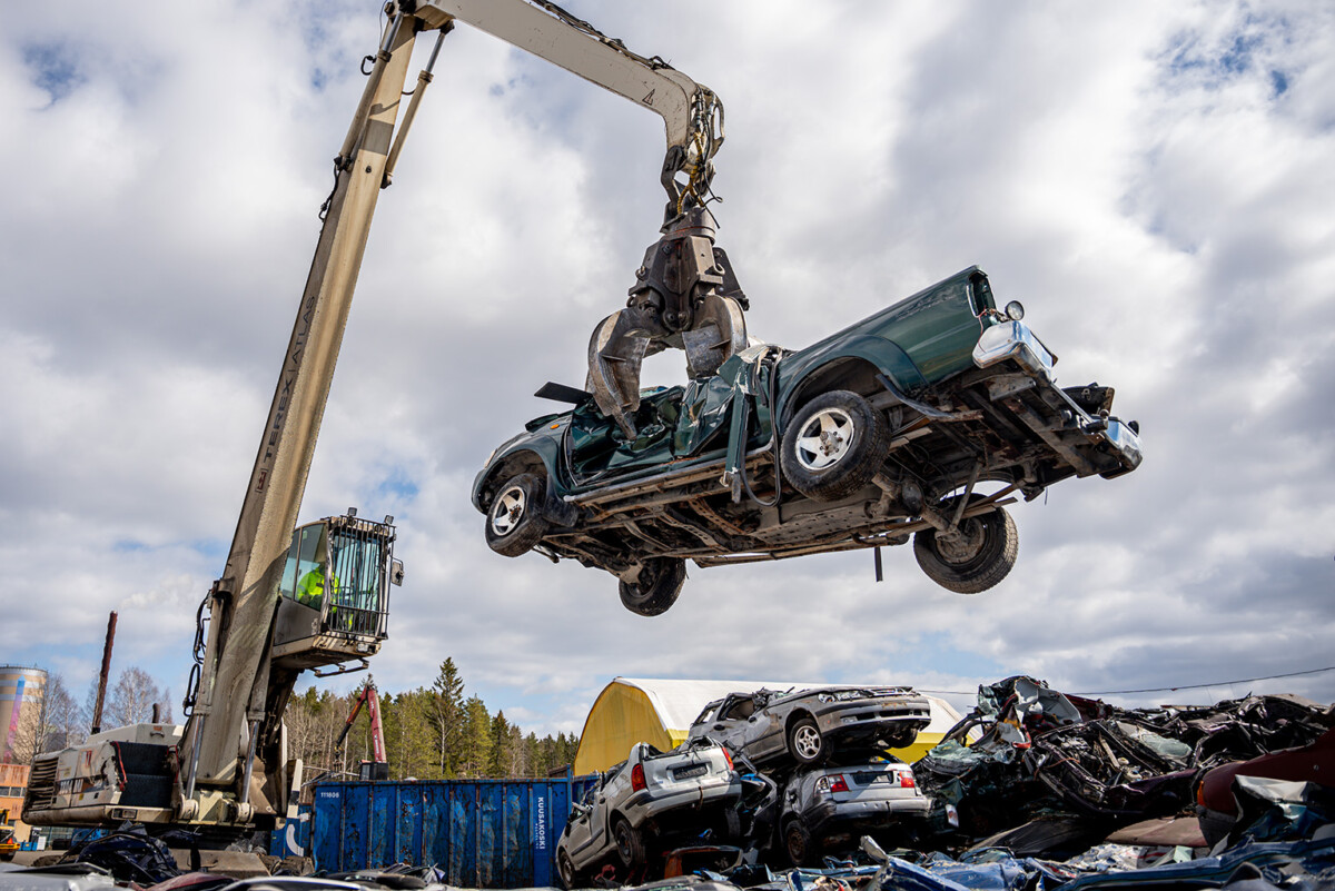 Suuri nosturi nostaa vaurioitunutta vihreää autoa romuttamolla, jonka alla on murskattuja ajoneuvokasoja ja taustalla puita pilvisen taivaan alla.