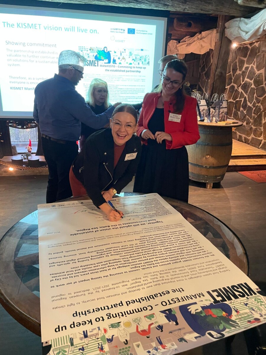 Hymyilevä nainen kumartuu pöydällä olevan suuren painetun asiakirjan yli ja allekirjoittaa sen, kun kolme muuta seisoo lähellä. Taustalla näkyy diaesitys KISMETin visio elää edelleen.