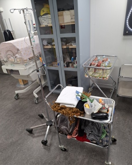 Sairaalahuone, jossa on tippateline, inkubaattori, metallikärry, jossa on pyyhkeitä, lääkintätarvikkeita ja peruukki, sekä vauvansänky, jossa on vauvanukkeja. Taustalla on kaappeja, joissa on hoitotarvikkeita.