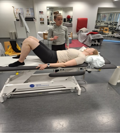 Nainen makaa hoitopöydällä polvet koukussa, kun terapeutti seisoo lähellä ja antaa ohjeita fysioterapian klinikalla.