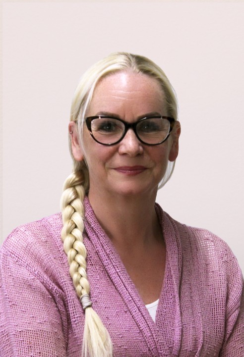 Nainen, jolla on pitkät vaaleat hiukset letissä, silmälasit ja vaaleanpunainen kuvioitu pusero, hymyilee lempeästi istuen vaalean taustan edessä.