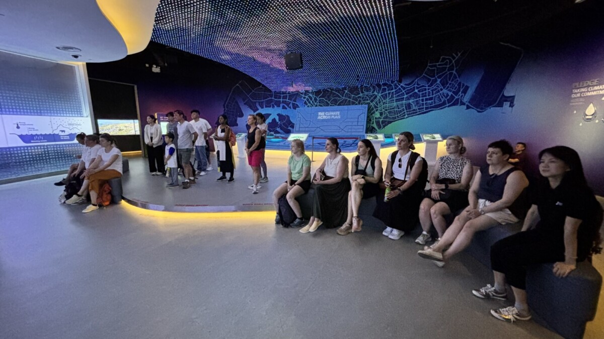 Joukko ihmisiä, sekä istuvia että seisovia, seuraa tarkkaavaisesti esitystä modernissa, hämärässä näyttelytilassa, jossa on sininen valaistus ja digitaaliset näytöt seinällä.