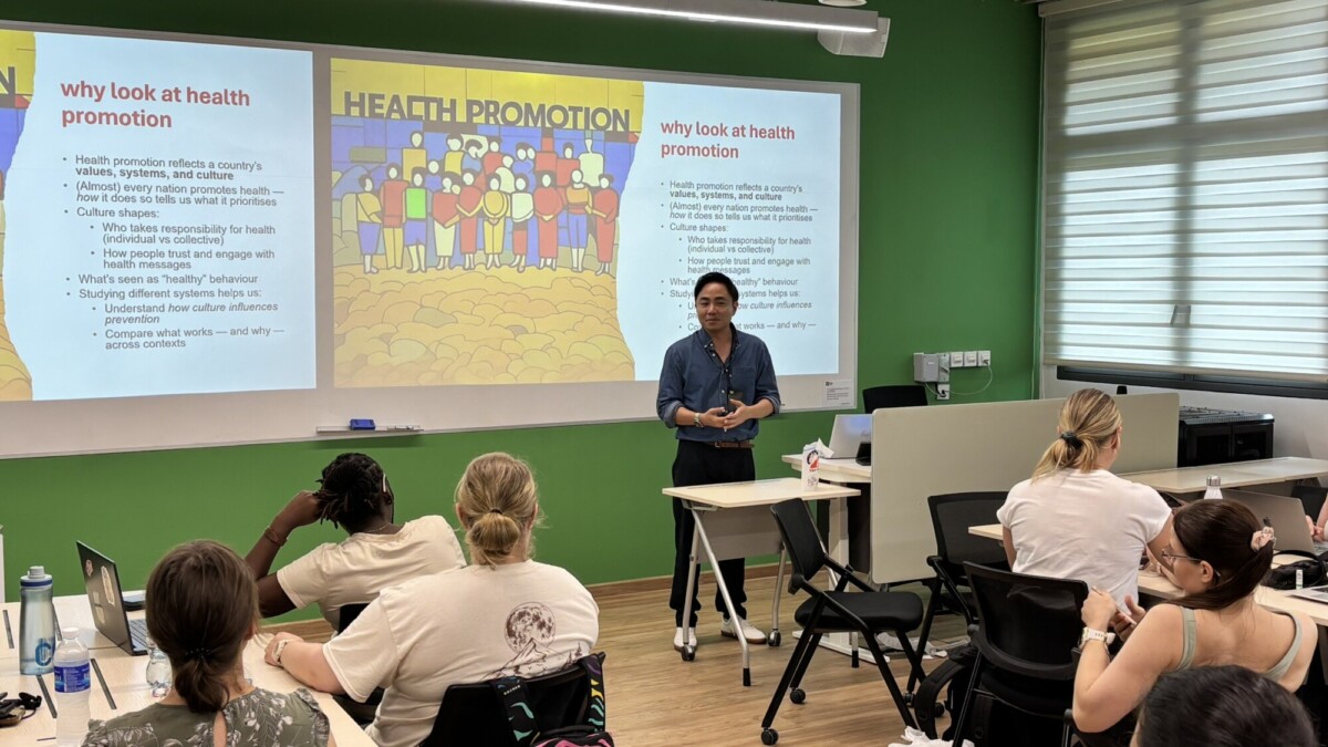 Esitelmöitsijä seisoo luokkahuoneen edessä, jossa on opiskelijoita, ja pitää luentoa terveyden edistämisestä. Hänen takanaan olevalle näytölle heijastetaan dioja otsikolla Why look at health promotion.