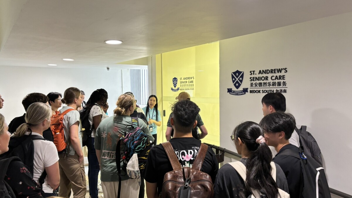 Joukko ihmisiä seisoo käytävällä ja kuuntelee opasta lähellä Bedok Southissa sijaitsevan St Andrew's Senior Care -hoitokodin sisäänkäyntiä. Seinällä näkyy kylttejä, joissa on keskuksen nimi ja logo.