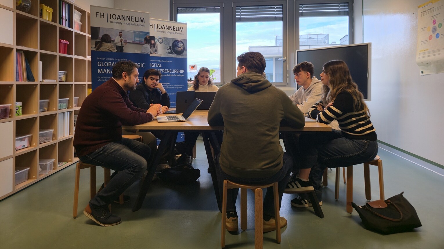 Seitsemän ihmistä istuu luokkahuoneessa pöydän ääressä keskustelemassa kannettavat tietokoneet auki. Taustalla näkyy bannereita ja kirjahyllyjä, ja suuret ikkunat päästävät luonnonvaloa sisään.