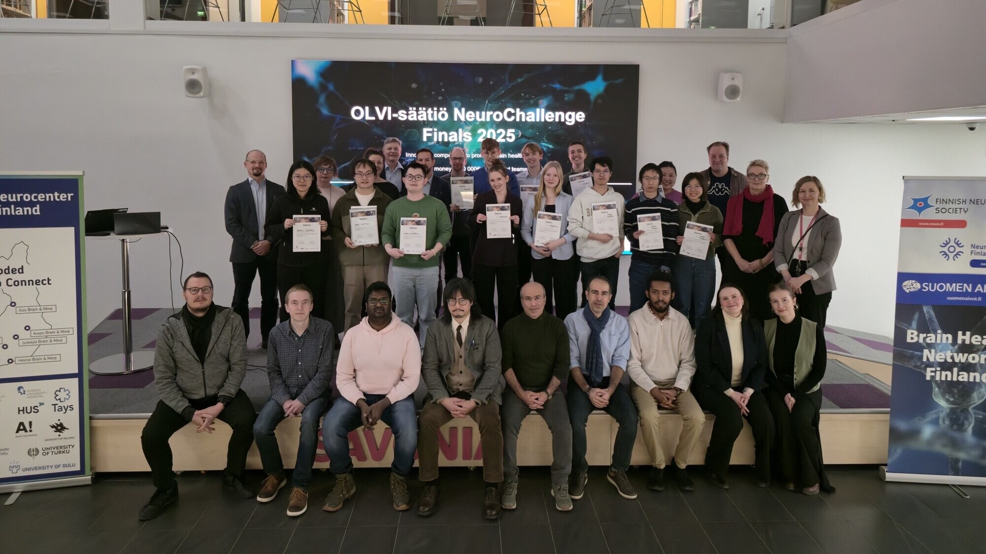 Joukko ihmisiä poseeraa sisätiloissa portailla ja näyttämön edessä, joillakin on kädessään todistuksia. Heidän takanaan suurella näytöllä lukee "OLVI-säätiö NeuroChallenge Finals 2025". Neurotieteestä kertovat banderollit ovat molemmin puolin.