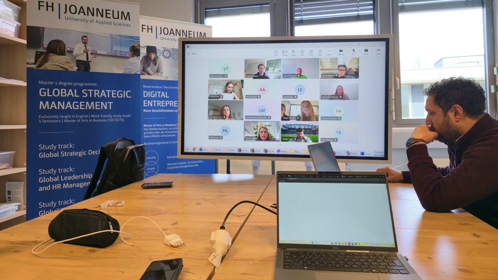 Mies istuu neuvottelupöydän ääressä kannettavien tietokoneiden kanssa ja osallistuu virtuaalikokoukseen, joka näkyy suurella näytöllä. Taustalla on FH Joanneumin globaalin strategisen johtamisen ja digitaalisen yrittäjyyden maisteriohjelmien bannereita.