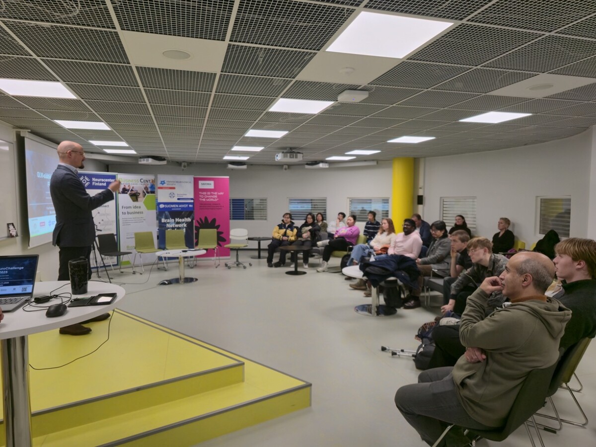 Pukumies esittelee modernissa kokoushuoneessa yleisölle. Osallistujat istuvat ja kuuntelevat tarkkaavaisesti; huoneen etuosassa on näkyvissä bannereita ja projektorinäyttö.