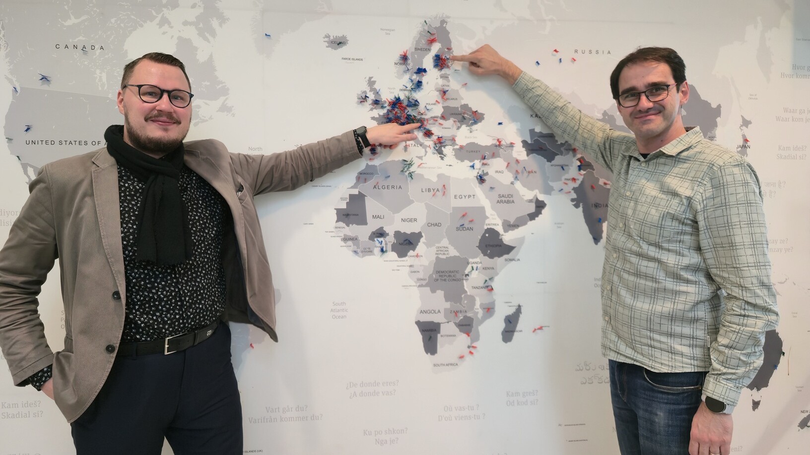 Kaksi miestä seisoo maailmankartan edessä, ja kumpikin osoittaa Eurooppaan. Vasemmalla olevalla miehellä on silmälasit, beige takki ja huivi, kun taas oikealla olevalla miehellä on silmälasit ja ruutupaita.