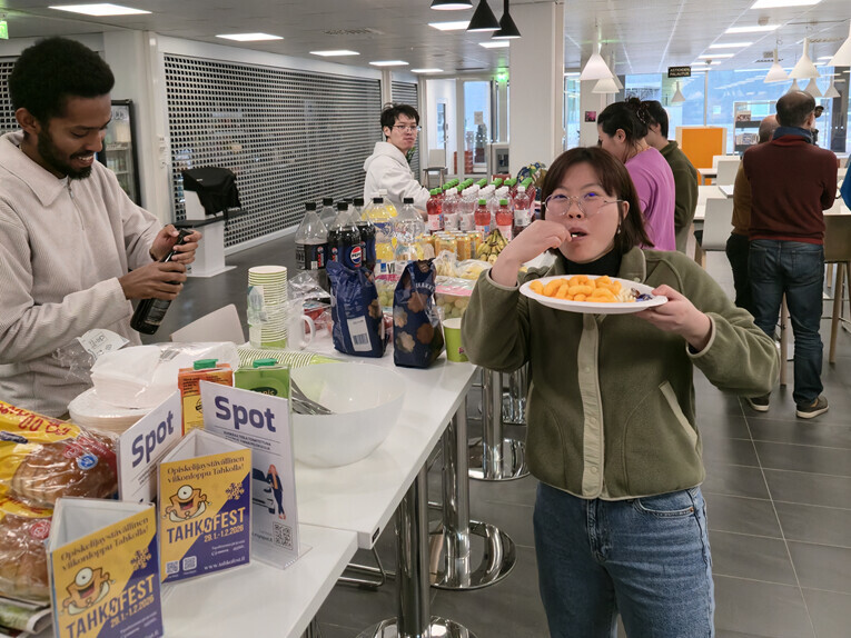 Henkilö seisoo etualalla syömässä lautaselta, kun muut kokoontuvat välipaloja ja juomia sisältävän pöydän ympärille modernissa, kirkkaasti valaistussa huoneessa. Jotkut ihmiset keskustelevat tai tarjoilevat itselleen ruokaa.