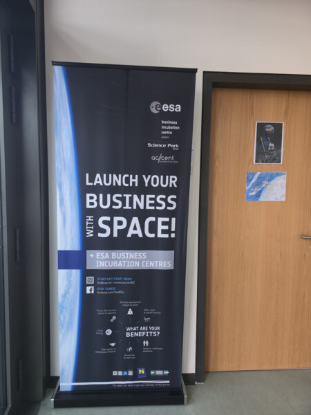 Oven vieressä olevassa pystysuorassa banderollissa lukee "Launch Your Business with Space!". ESA Business Incubation Centres", jossa on ESAn logoja ja tietoja ESA:sta, yhteistyökumppaneista ja startup-yrityksille tarjottavista eduista. Sen vieressä olevalla seinällä on pieni astronauttijuliste.