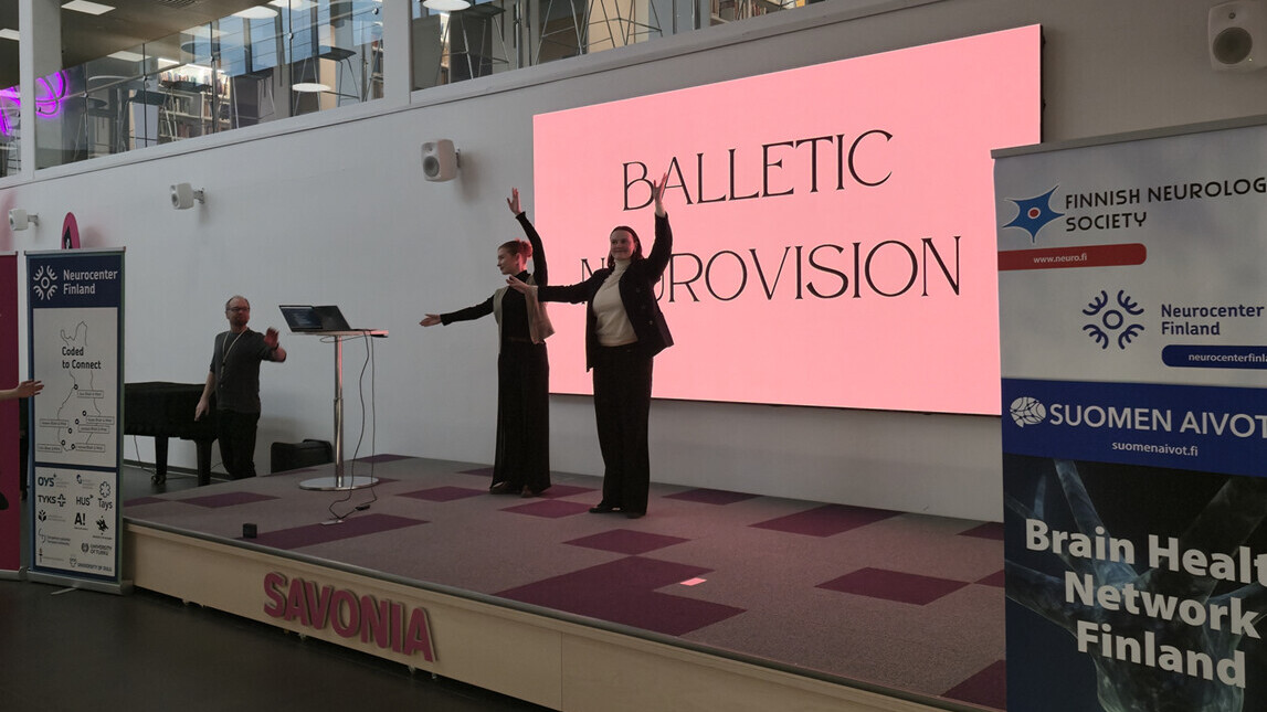 Kaksi esiintyjää seisoo lavalla kädet kohotettuna suuren näytön edessä, jossa lukee BALLETIC IMPROVISION. Vasemmalla soittaa pianisti. Lavalla on esillä neurologisten ja aivoterveysjärjestöjen banderolleja.