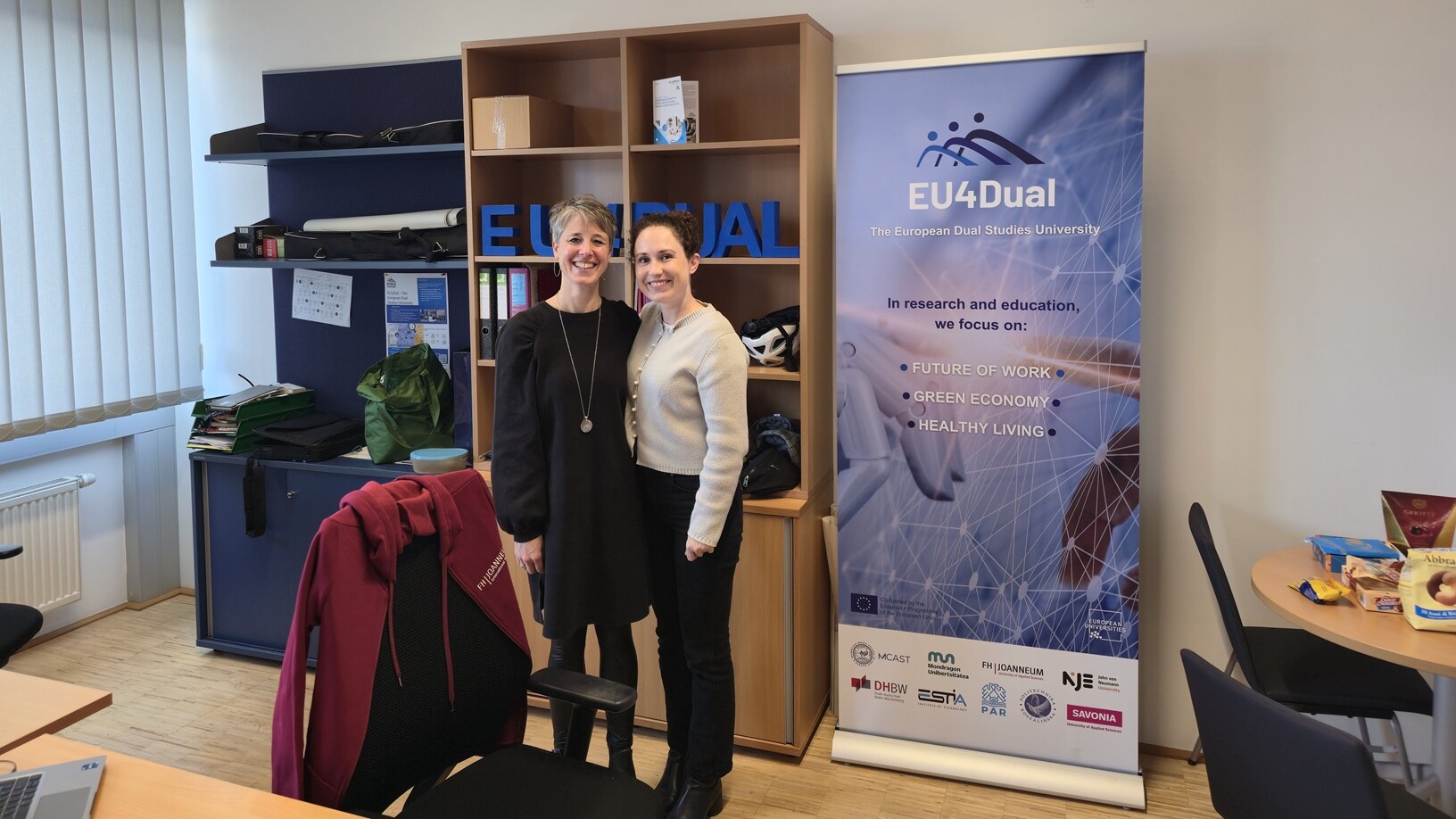 Kaksi naista seisoo hymyillen toimistossa sinisen EU4Dual-banderollin vieressä. Taustalla näkyy hyllyjä, joissa on kirjoja ja kansioita, ikkuna, jossa on kaihtimet, ja pyöreä pöytä, jossa on papereita ja punainen takki.
