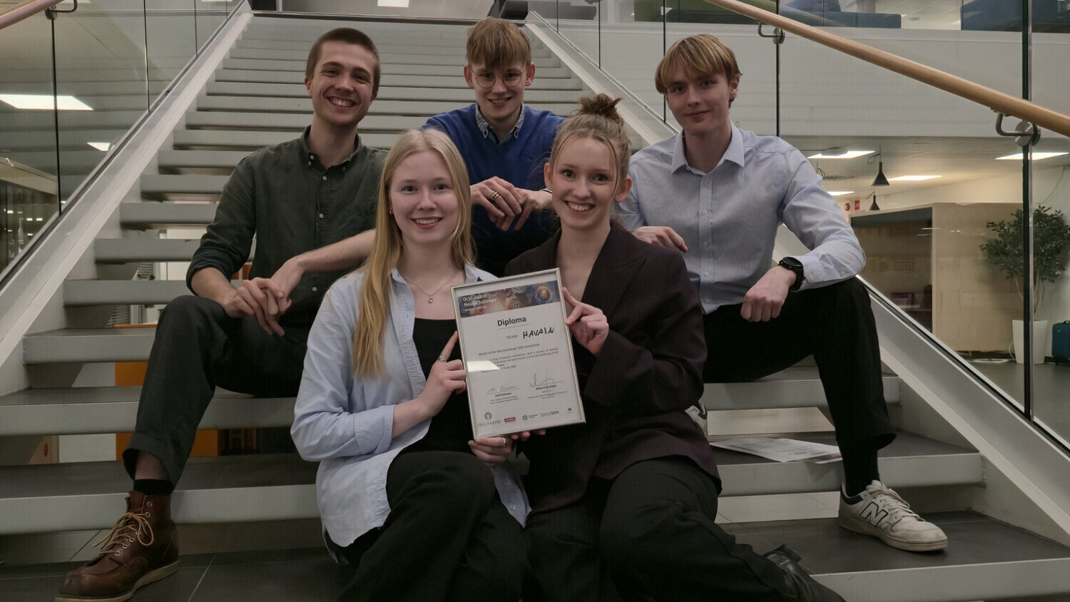 Viisi hymyilevää nuorta aikuista istuu ja poseeraa sisätiloissa portailla, kaksi edessä pitelee todistusta, ja taustalla on moderni sisustus ja vaaleanpunaisia neonkylttejä.