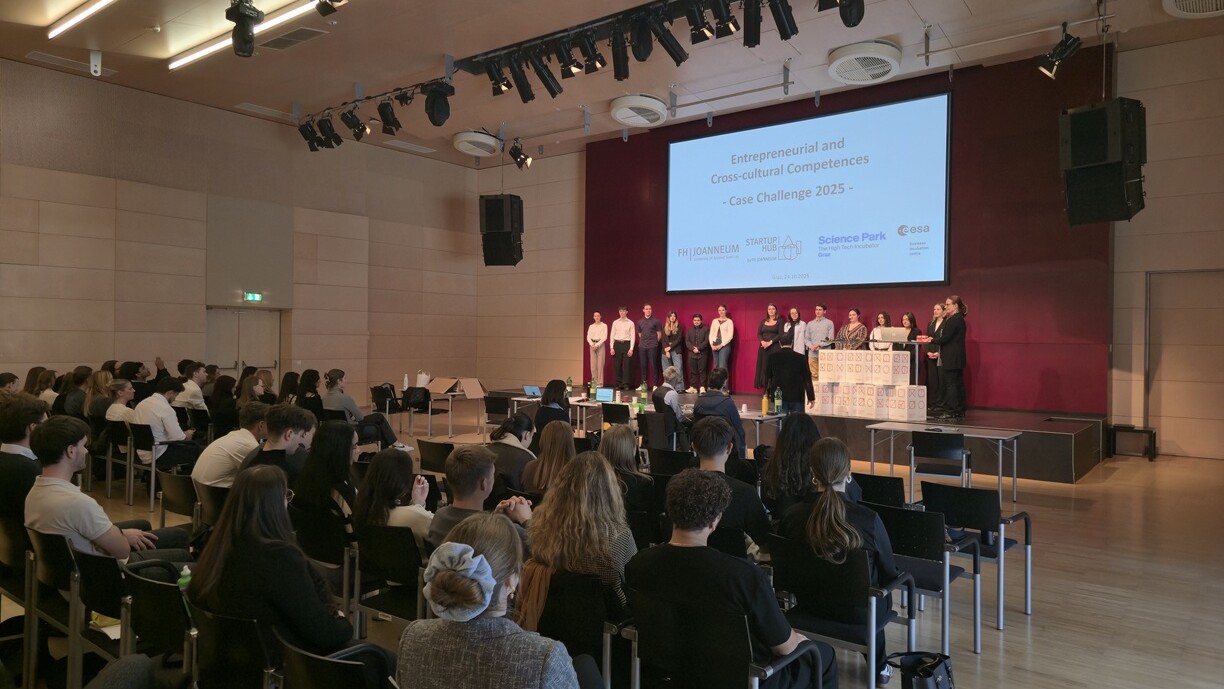 Suuri joukko ihmisiä istuu riveissä lavaa vastapäätä, jossa useat henkilöt seisovat näytön vieressä, jossa näkyy "Yrittäjyys- ja kulttuurienvälinen osaaminen - Case Challenge 2025". Yleisö seuraa tarkkaavaisena modernissa salissa.