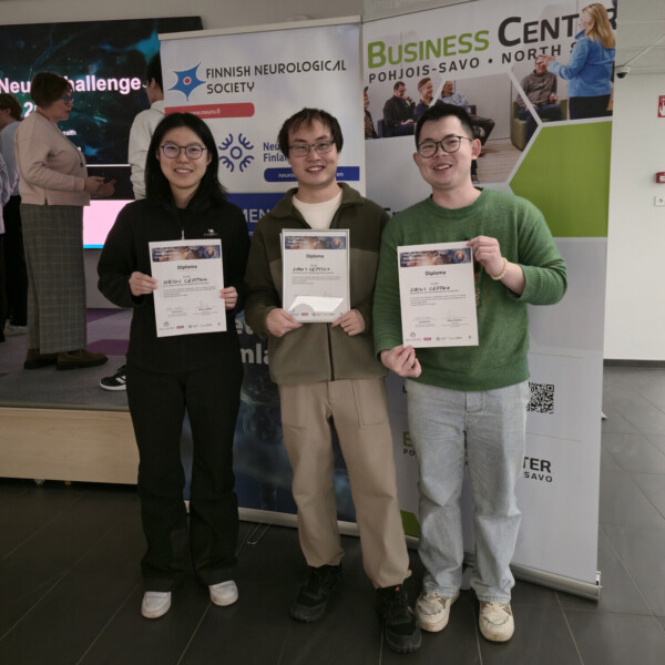 Kolme ihmistä seisoo sisätiloissa hymyillen ja todistukset kädessään Suomen Neurologiyhdistyksen ja Liikekeskus Pohjois-Savon banderollien edessä. Taustalla näkyy muita ihmisiä ja lava.