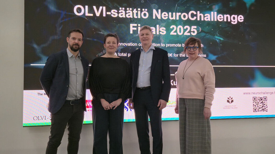 Neljä ihmistä seisoo ja poseeraa suuren näytön edessä, jossa lukee teksti OLVI-säätiö NeuroChallenge Finals 2025. Ryhmä hymyilee modernissa sisätilassa, jossa on violetti ja harmaa matto.