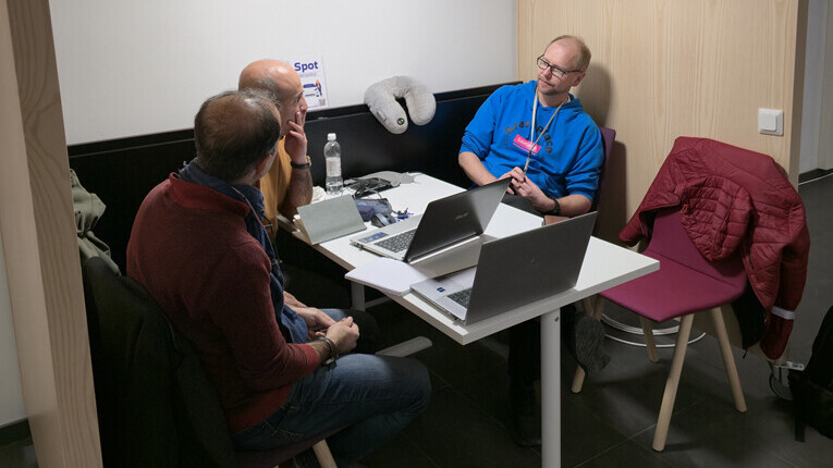 Kolme miestä istuu pienen pöydän ääressä modernissa kopissa keskustelemassa. Pöydällä on kaksi kannettavaa tietokonetta, papereita ja vesipullo. Takit on ripustettu lähistöllä oleville tyhjille tuoleille. Ympäristö vaikuttaa rennolta kokoustilalta.