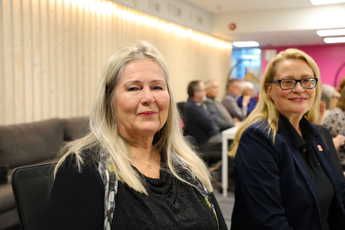 Kaksi vanhempaa naista, joilla on pitkät vaaleat hiukset, istuvat sisätiloissa tapahtumassa, katsovat kameraan ja hymyilevät. Taustalla istuu useita muita ihmisiä, ja huoneessa on vaaleat seinät ja kirkas valaistus.