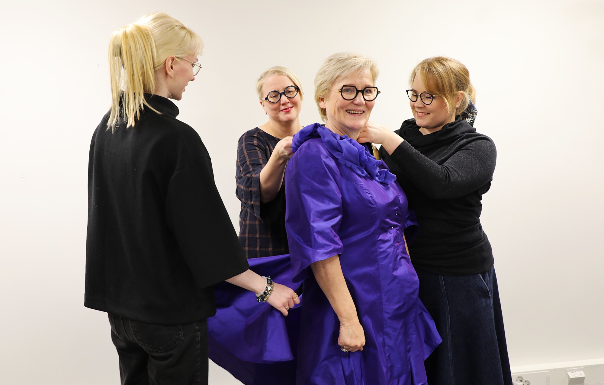 Neljä naista seisoo yhdessä hymyillen. Kolme heistä säätää kirkkaan violettia, röyhelöistä mekkoa, jota keskellä oleva vanhempi nainen käyttää. Tausta on pelkkä valkoinen, ja ryhmä vaikuttaa iloiselta ja yhteistyökykyiseltä.