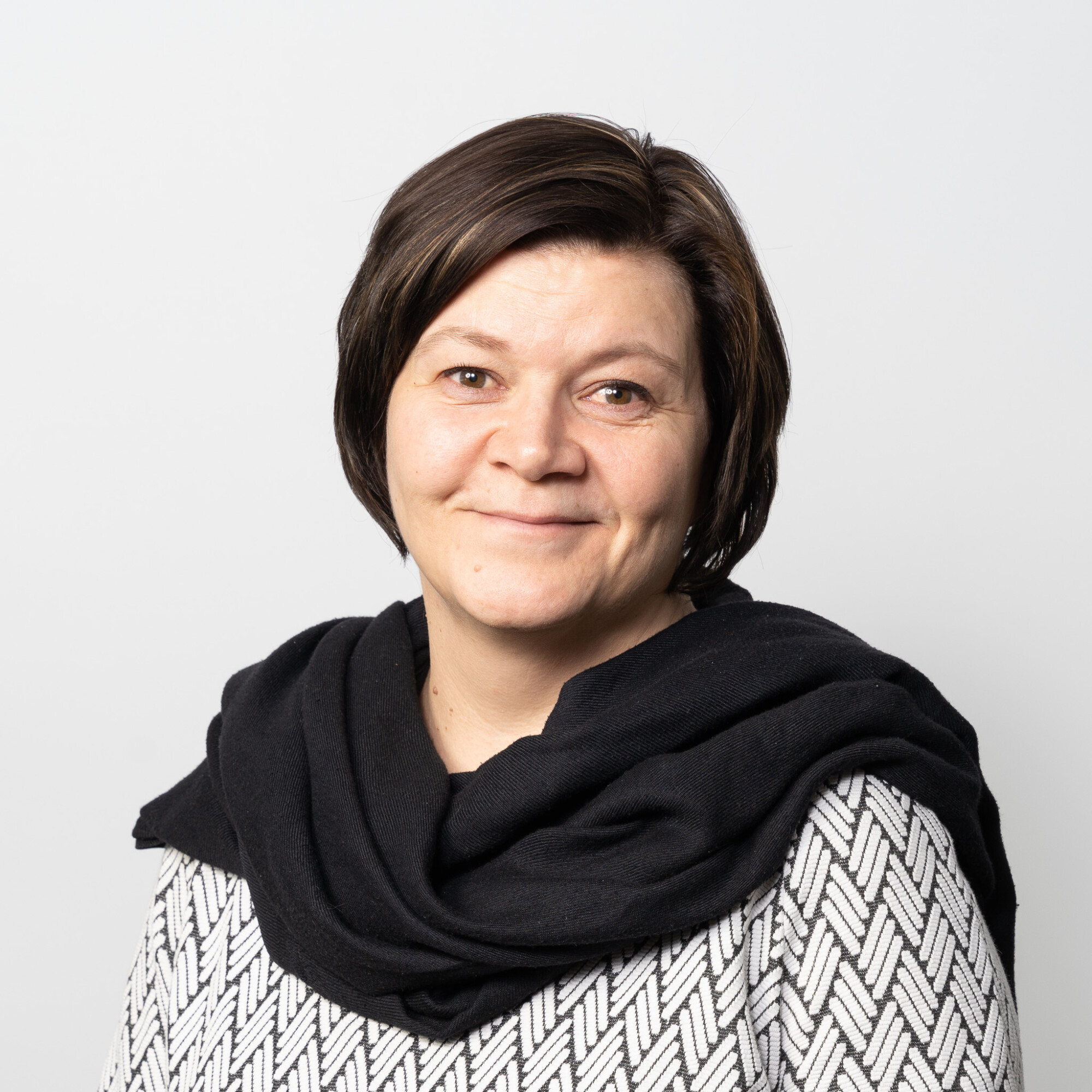 Lyhyet ruskeat hiukset ja hento hymy kasvoillaan kameraan. Hänellä on päällään kuviollinen villapaita ja hartioilleen kiedottu musta huivi, ja hän seisoo vaaleaa taustaa vasten.