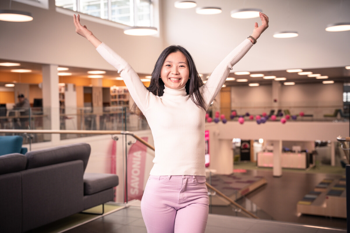 Hymyilevä nainen, jolla on valkoinen poolokaulus ja vaaleanvioletit housut, seisoo sisätiloissa kädet ylhäällä. Hän on modernissa, hyvin valaistussa rakennuksessa, jonka taustalla on sohvia ja avointa tilaa.