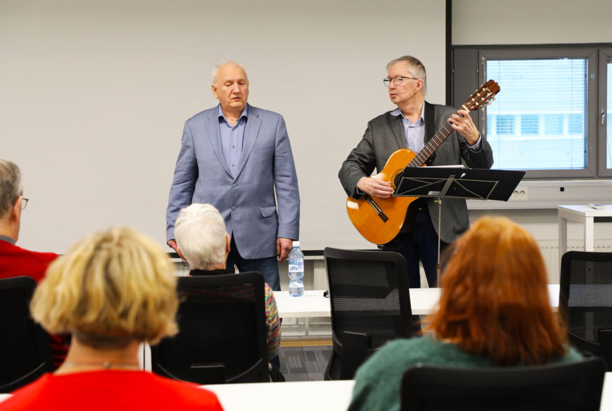 Kaksi vanhempaa miestä esiintyy luokkahuoneessa yleisön edessä; toinen mies laulaa ja toinen soittaa akustista kitaraa. Molemmilla on bleiserit, ja useita ihmisiä istuu heitä vastapäätä. Heidän takanaan näkyy ikkuna ja valkokangas.