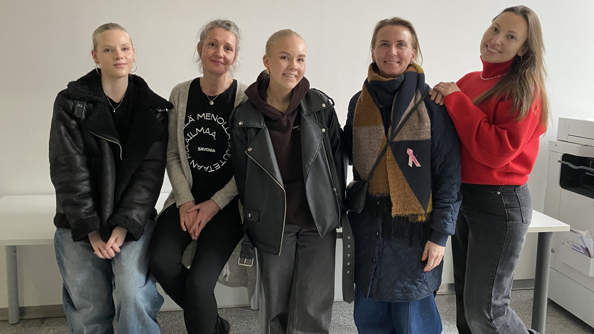 Viisi naista seisoo ja istuu yhdessä sisätiloissa hymyillen kameralle. Heillä on rennot ja talvivaatteet, kuten takit, huivit ja villapaidat. Taustalla on tavallinen valkoinen seinä, jossa on toimistokalusteita.