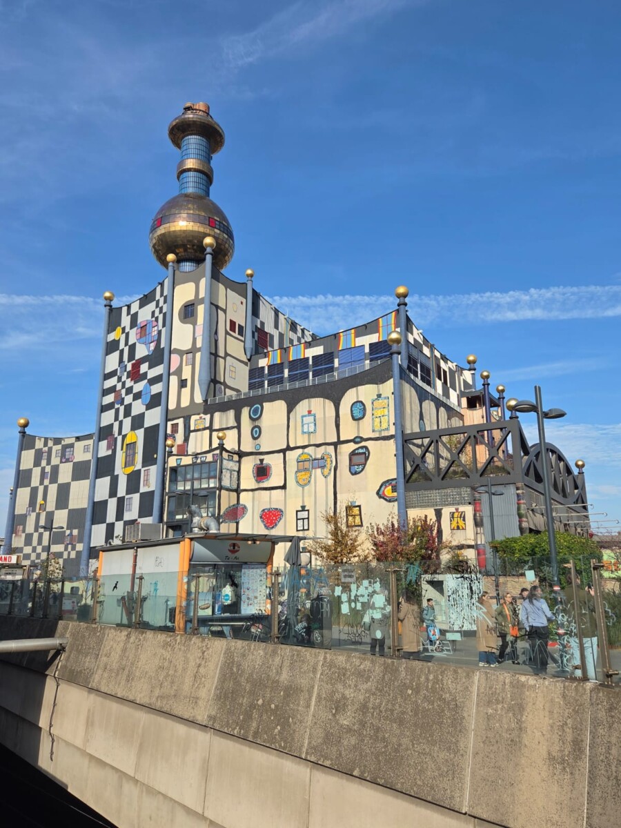 Värikäs, moderni rakennus, jossa on ruudullinen julkisivu, omituisia muotoja ja suuri kultainen pallo tornin huipulla kirkkaalla sinisellä taivaalla. Alueen edessä on lasipaneeleita ja viherkasveja.