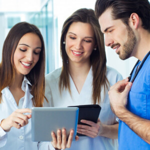 Kolme nuorta terveydenhuollon ammattilaista, kaksi naista ja yksi mies, seisovat yhdessä hymyillen ja keskustelevat jostain digitaalisella tabletilla. Miehellä on siniset leikkaushaalarit ja stetoskooppi, naisilla valkoiset takit.