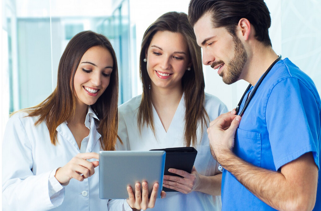 Kolme nuorta terveydenhuollon ammattilaista, kaksi naista ja yksi mies, seisovat yhdessä hymyillen ja keskustelevat jostain digitaalisella tabletilla. Miehellä on siniset leikkaushaalarit ja stetoskooppi, naisilla valkoiset takit.