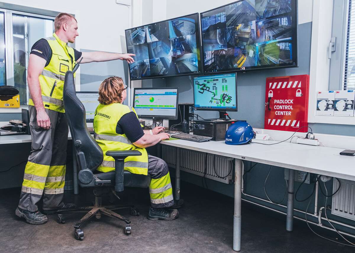 Kaksi turvaliiveihin pukeutunutta työntekijää tarkkailee useita valvontakameroita ja tietokonenäyttöjä valvomossa, jonka nimi on Padlock Control Center. Toinen henkilö istuu, kun taas toinen seisoo ja osoittaa näyttöä.