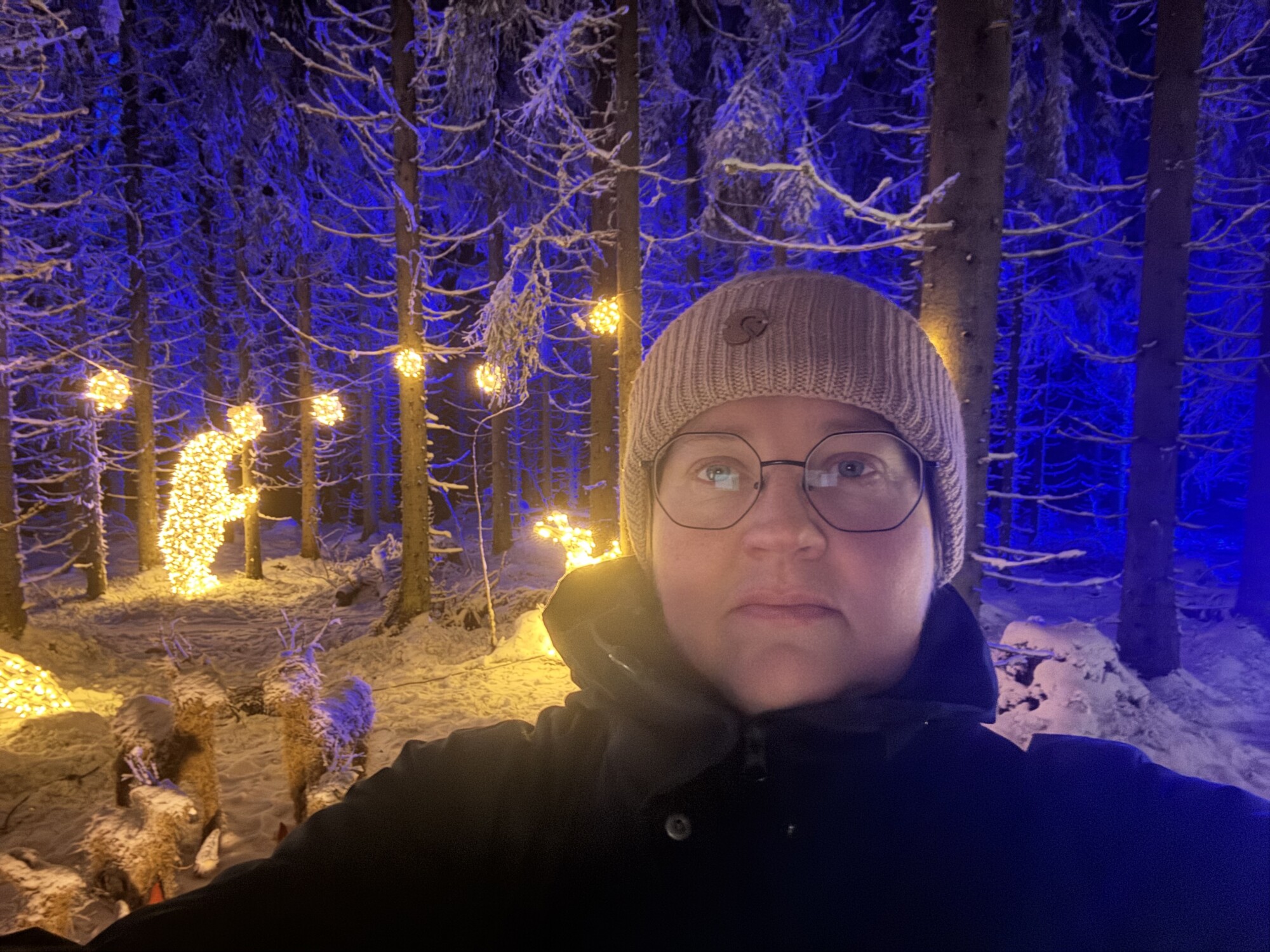 Silmälasit ja pipo päässä oleva henkilö ottaa selfietä yöllisessä lumisessa metsässä, jossa puut ja eläimenmuotoiset jouluvalot hehkuvat lämpimästi syvänsinistä taustaa vasten.