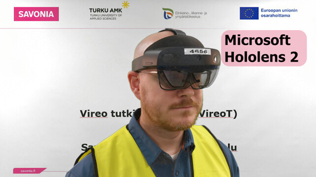 Henkilö, jolla on Microsoft Hololens 2 -kuulokkeet ja keltainen turvaliivi, seisoo näytön edessä, jossa näkyy erilaisia logoja ja tekstejä, kuten Savonia ja Microsoft Hololens 2.