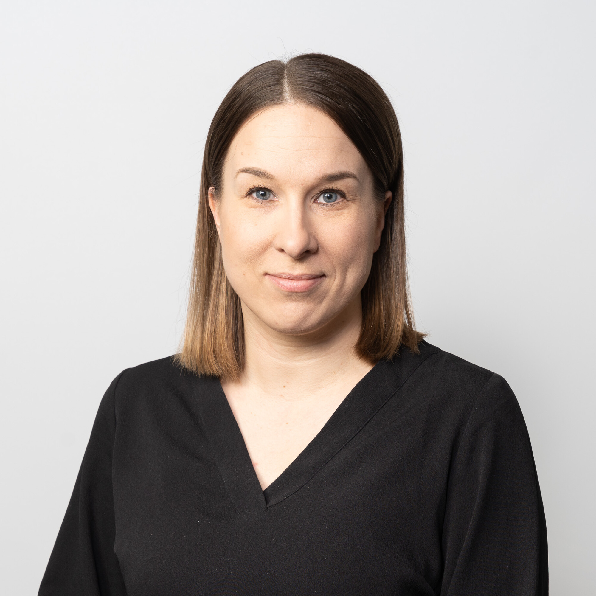 Naisella, jolla on suorat, olkapään pituiset ruskeat hiukset ja vaalea iho, on yllään musta V-kaula-aukkoinen toppi ja hän hymyilee kameralle pehmeästi vaaleanharmaalla taustalla.