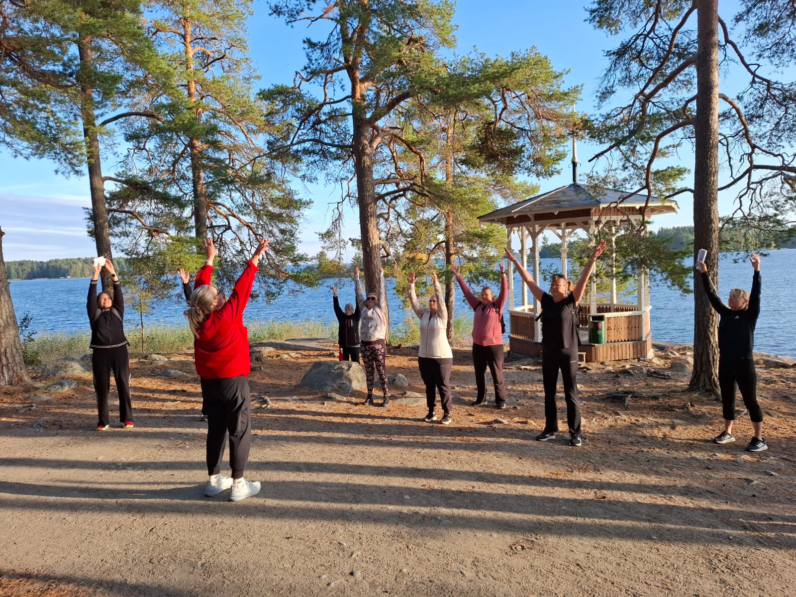 Ryhmä ihmisiä harjoittelee ulkona hiekkatiellä järven lähellä, korkeiden mäntyjen ympäröimänä. He venyttelevät kädet kohotettuna punatakkisen ohjaajan johdolla. Taustalla näkyy pieni huvimaja ja järvi.