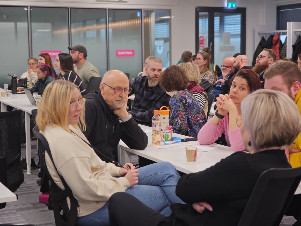 Joukko aikuisia istuu ryhmittäin pöydissä modernissa kokoushuoneessa ja keskustelee vilkkaasti. Jotkut kuuntelevat, toiset puhuvat. Ilmapiiri vaikuttaa yhteistyökykyiseltä ja keskittyneeltä.