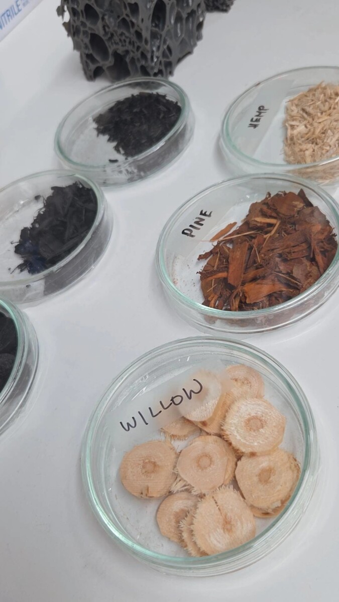 Kuusi lasiastiaa, joissa on merkittyjä näytteitä eri puumateriaaleista - puuhiiltä, mäntyä, hamppua ja pajua - on esillä valkoisella pinnalla. Kukin astia sisältää erityyppistä ja -rakenteista kasvimateriaalia.