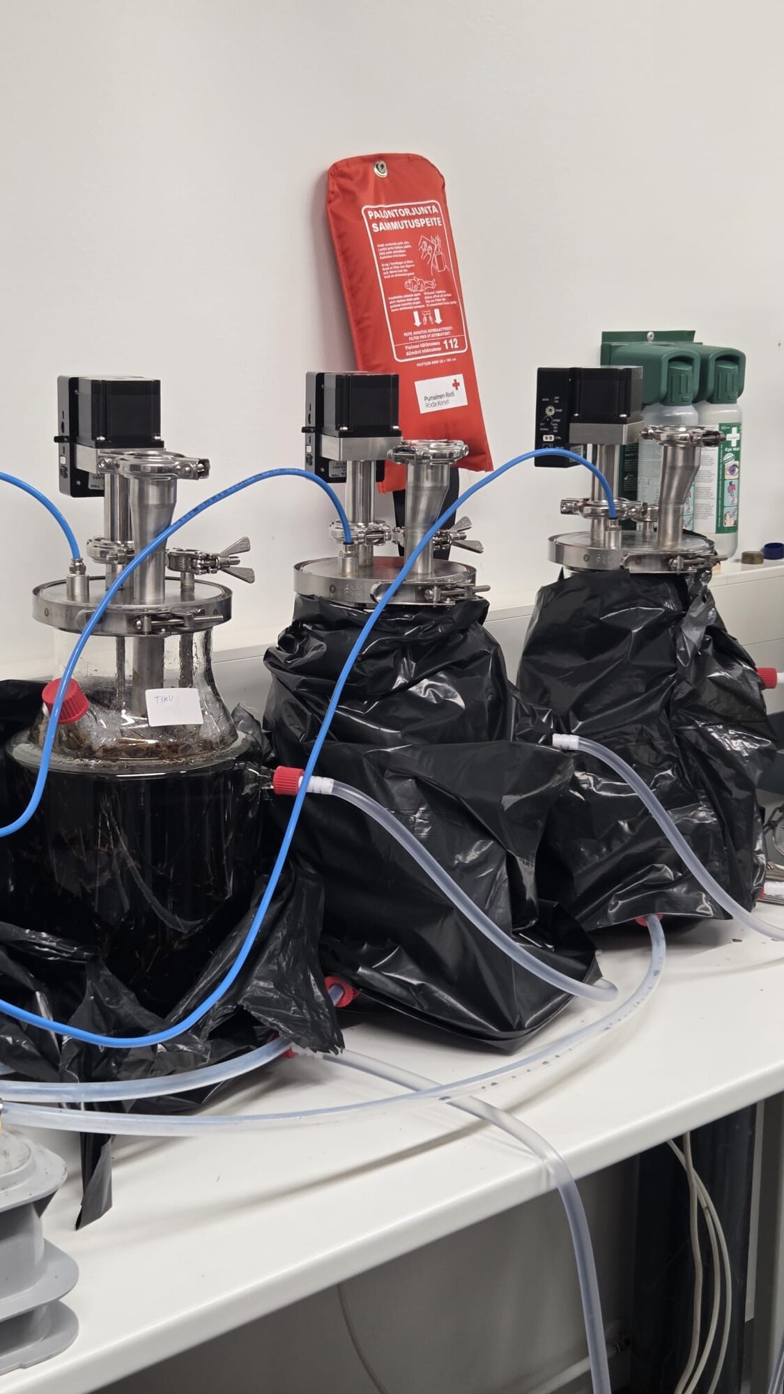 Kolme mustiin muovipusseihin käärittyä bioreaktoria on yhdistetty sinisillä ja punaisilla letkuilla laboratoriopöydällä. Niiden takana olevalle valkoiselle seinälle on asennettu punainen turvapakkaus ja vihreät laitteet. Laboratoriolaitteet ovat lähellä.