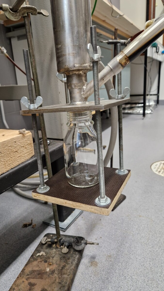 Lasipulloa pidetään jalustalla laboratoriolaitteiden alla, ja sen yläpuolella on metallilaite ja letkut, todennäköisesti nesteen tai kaasun keräämistä varten laboratorioympäristössä. Asetelma on harmaalla lattialla, jossa on erilaisia laboratoriomateriaaleja.