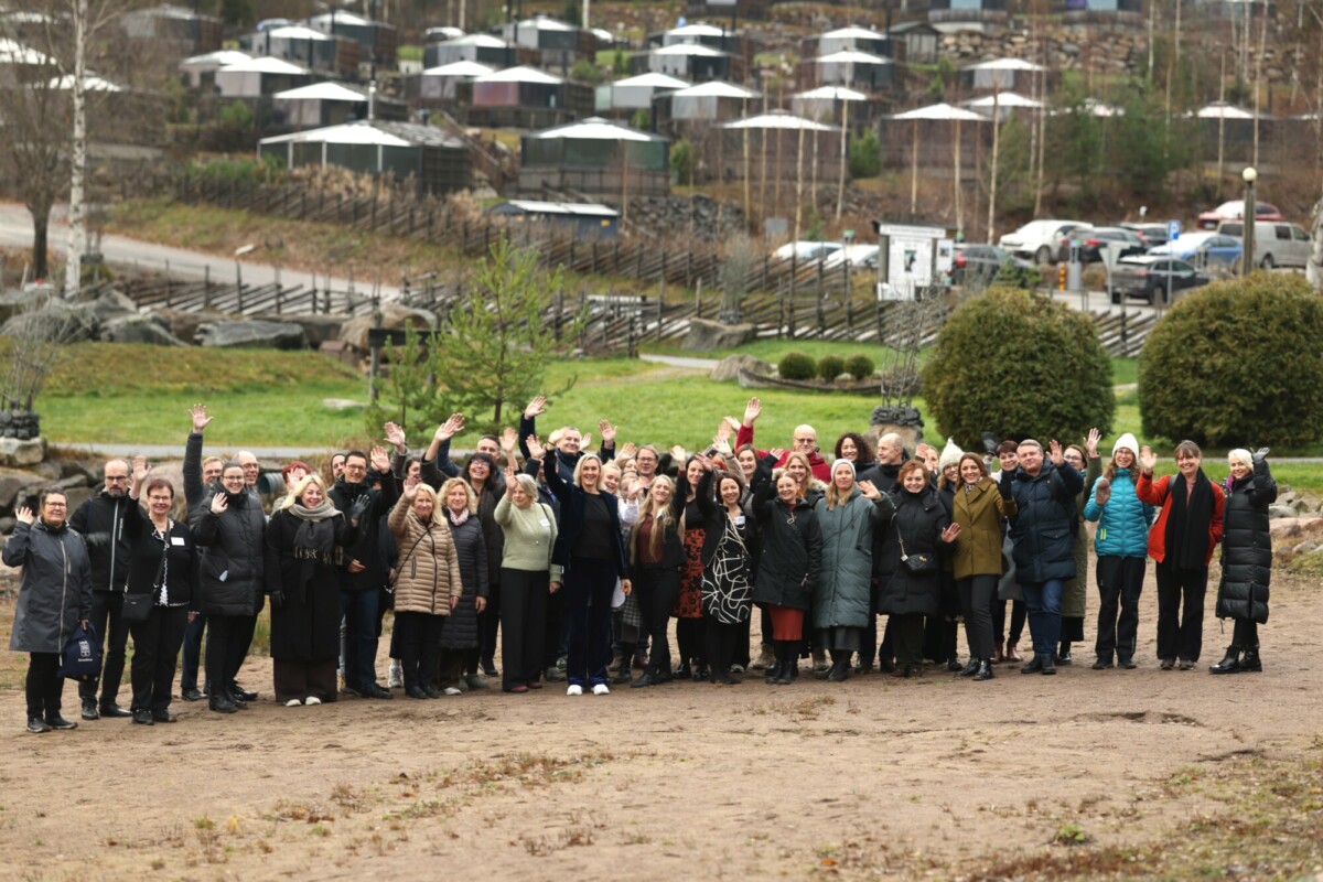 Suuri joukko ihmisiä seisoo ulkona hiekkatiellä hymyillen ja vilkuttaen kameralle. Heidän takanaan on puita, pensaita ja useita pieniä mökkejä tai majoja, joissa on valkoiset katot. Tunnelma näyttää iloiselta ja rennolta.
