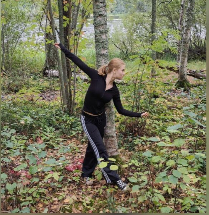 Mustiin urheiluvaatteisiin pukeutunut nainen poseeraa toinen käsi kohotettuna ja toinen ojennettuna metsässä, jota ympäröi vehreys ja puut, ja taustalla näkyy vesistö.