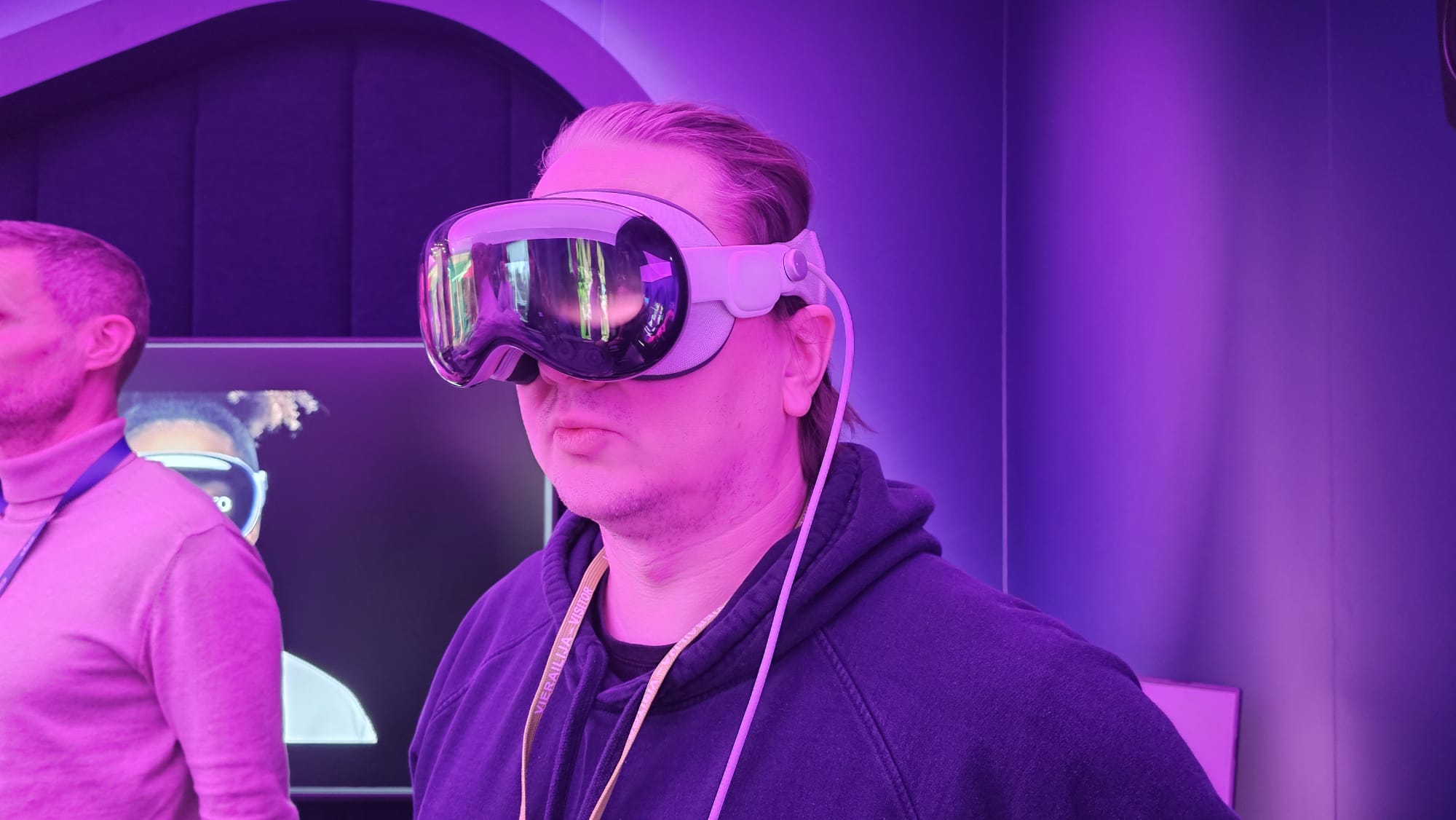 Henkilö, jolla on virtuaalitodellisuuskuulokkeet, seisoo huoneessa, jossa on violetti valaistus. Taustalla näkyy toinen henkilö, ja näytöllä näkyy kuva henkilöstä, jolla on myös VR-kuulokkeet.