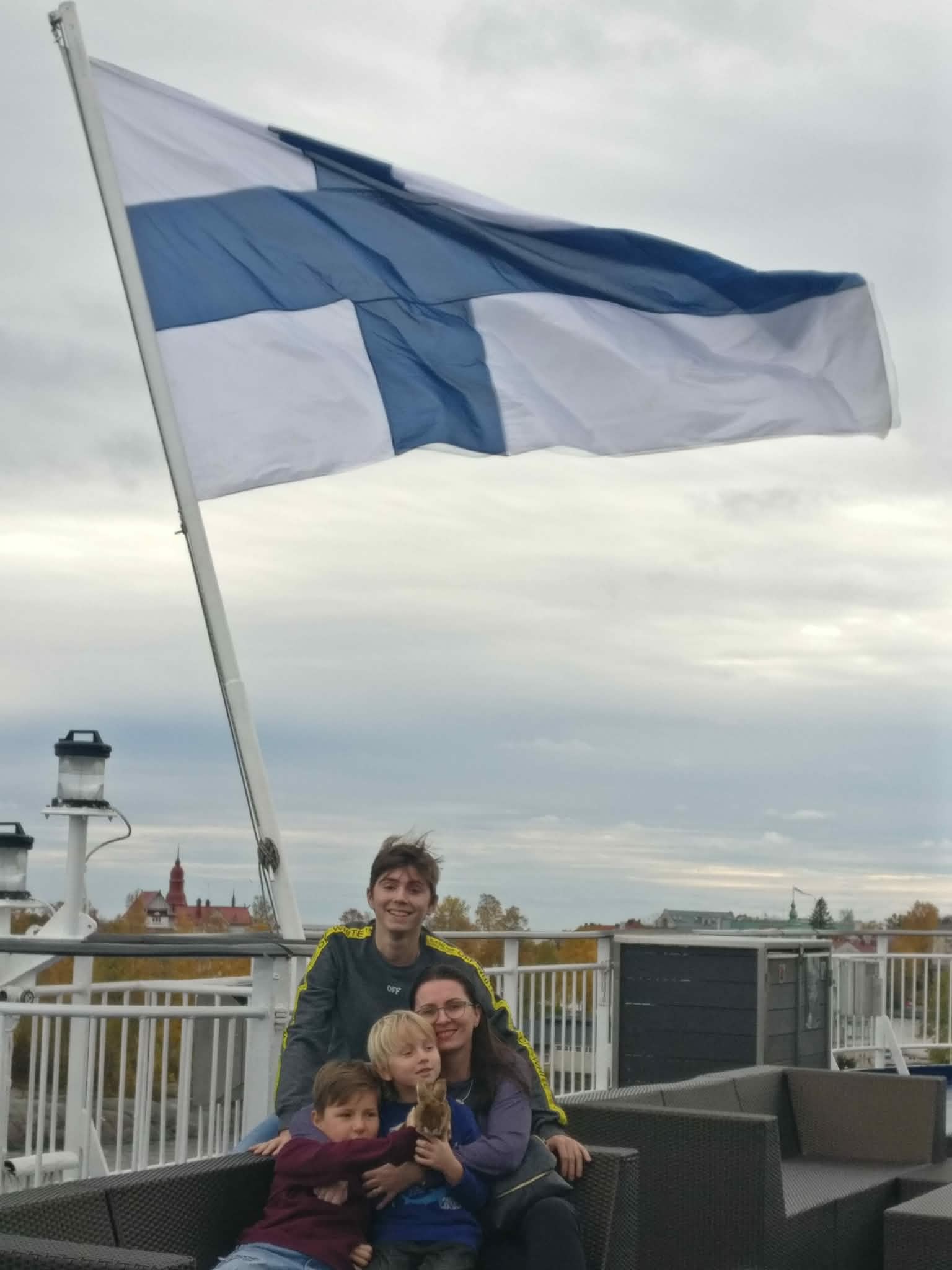 Nelihenkinen perhe istuu hymyillen ulkokalusteilla, ja taustalla liehuu suuri Suomen lippu. Taivas on pilvinen, ja kaukaisuudessa näkyy joitakin rakennuksia ja puita.