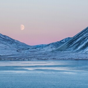 Lumipeitteiset vuoret ympäröivät jäätynyttä järveä pastellivärisen taivaan alla hämärässä, ja puolikuu näkyy horisontin yläpuolella.