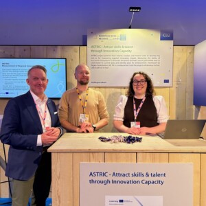 Kolme ihmistä seisoo puisen kopin takana tapahtumassa ja hymyilee kameralle. Kojussa on ASTRICin kylttejä, tiskillä on kannettava tietokone ja niiden takana on tiedotusjulisteita.