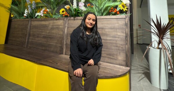 Nainen, jolla on pitkät tummat hiukset, istuu puisella penkillä erilaisilla vihreillä kasveilla ja värikkäillä kukilla koristellun istutusruukun edessä kirkkaasti valaistussa sisätilassa, jossa on keltaisia korostuksia.